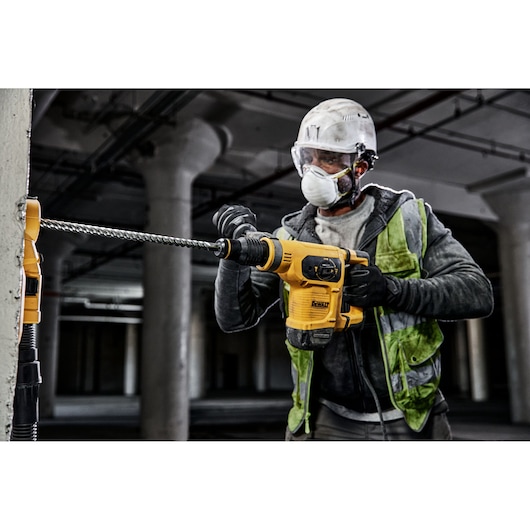 A construction worker wearing a safety helmet and vest is using a yellow DEWALT rotary hammer drill to drill into a concrete