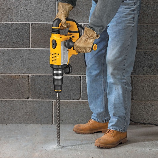 A person using a yellow DEWALT rotary hammer drill with a long drill bit to drill into a concrete floor, standing next to a