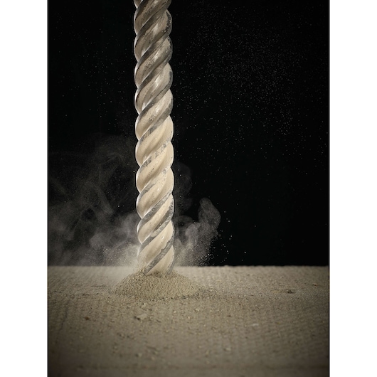 Close-up view of a DEWALT spiral masonry drill bit drilling into a concrete surface, with dust and debris being released.