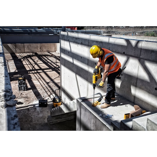 A construction worker wearing a yellow hard hat and orange safety vest uses a DEWALT rotary hammer drill to drill into a