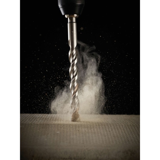 A close-up of a DEWALT drill bit in action, drilling into a concrete surface with dust rising around the bit.