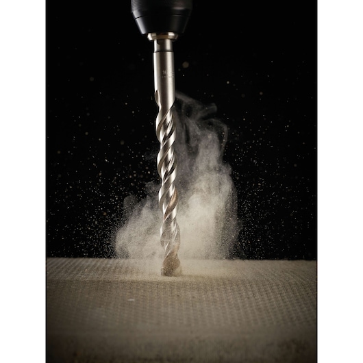 Close-up of a DEWALT metal drill bit attached to a drill, drilling into a concrete surface with dust spreading around.