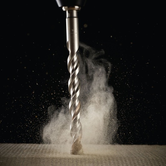 Close-up of a DEWALT drill bit cutting into material, dust rising, against a black background.