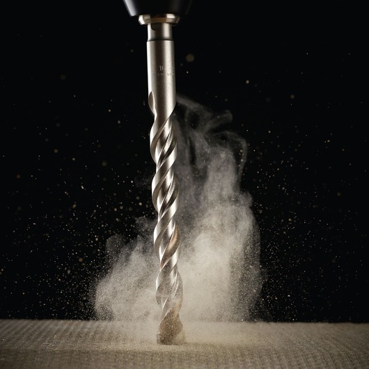 Metal drill bit drilling into a surface, with dust clouds rising up against a dark background.