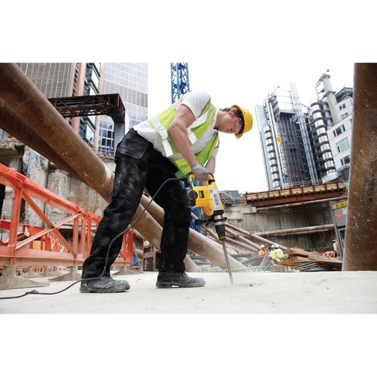 A construction worker wearing a safety vest and helmet uses a yellow DEWALT power tool, likely a hammer drill, to work on a