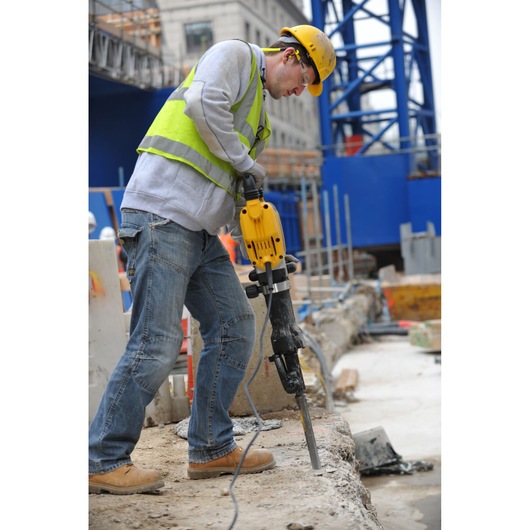 Worker using a yellow DEWALT jackhammer on concrete at a construction site, wearing helmet and safety vest.