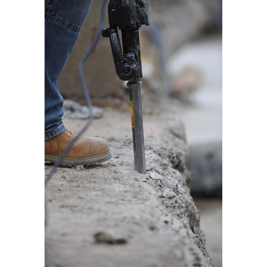 A close-up of a person using a DEWALT drill bit to break concrete. The person is wearing jeans and work boots.