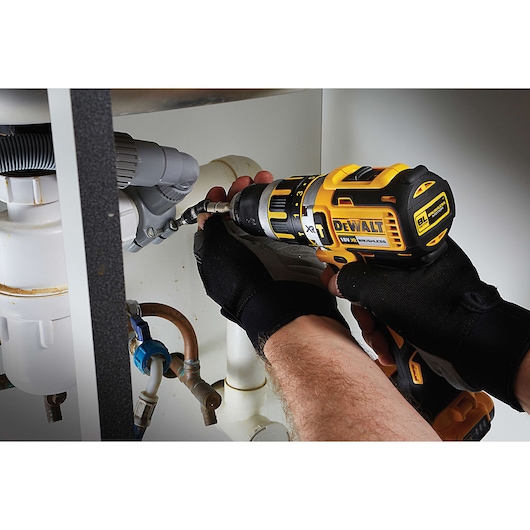 Close-up of a person using a DEWALT power drill to fasten a screw onto plumbing under a sink. The person is wearing black gloves.