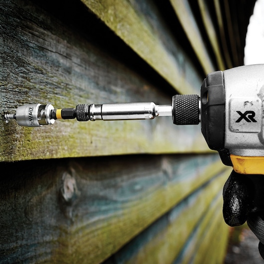A close-up of a DEWALT screwdriving bit and holder attached to a power tool, driving a screw into an exterior wooden wall.