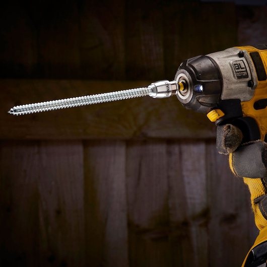 A DEWALT drill with a long screw attachment held by a gloved hand against a wooden background.