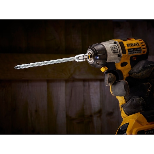 Gloved hand holding a DEWALT impact driver with a long screw attached, shown against a wooden background.