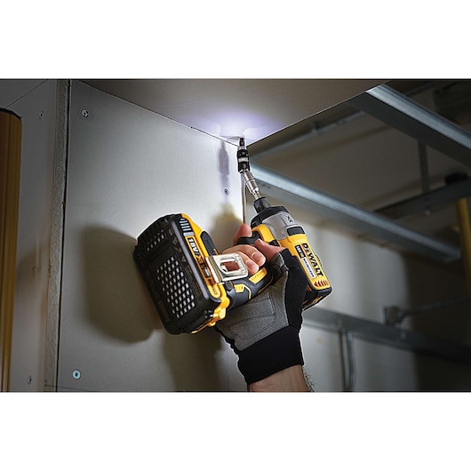 A person uses a DEWALT cordless drill to screw into a ceiling panel, wearing a protective glove. The drill is equipped with a bit and an LED light, illuminating the workspace.