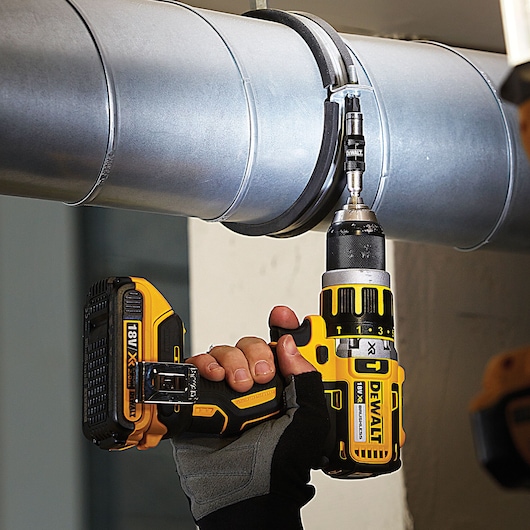 A person using a DEWALT 18V cordless drill to fasten a screw into a metal pipe. The drill is yellow and black and is held with a gloved hand.