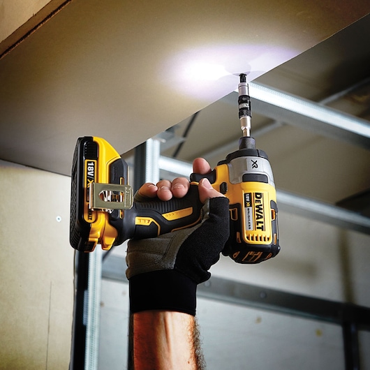 A person wearing a work glove uses a DEWALT 18V XR cordless impact driver with a drill bit to install screws into a ceiling. The tool emits an LED light to illuminate the work area.