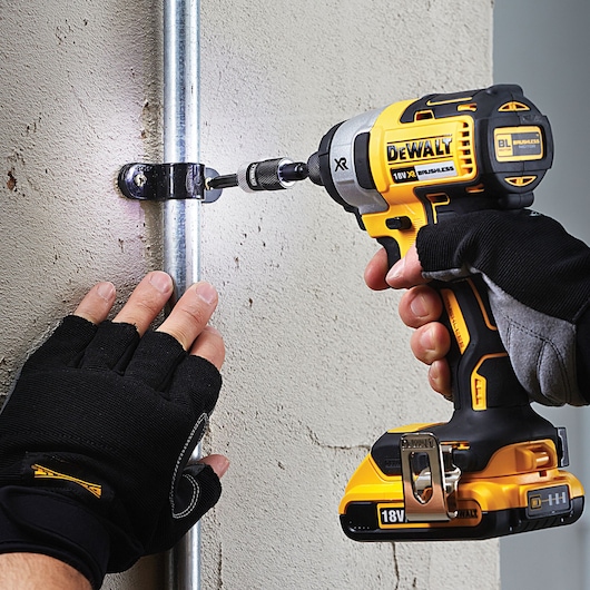 A person wearing gloves uses a DEWALT 18V XR brushless cordless impact driver to fasten a metal clamp onto a pipe mounted on a concrete wall.