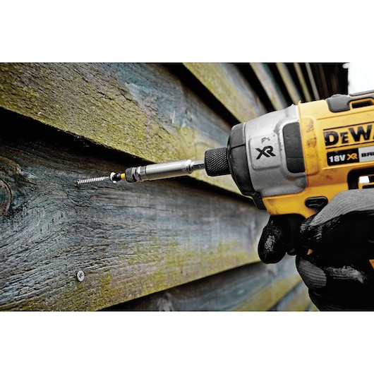 A DEWALT cordless power drill is being used to drive a screw into a wooden plank. The hand holding the drill is wearing a