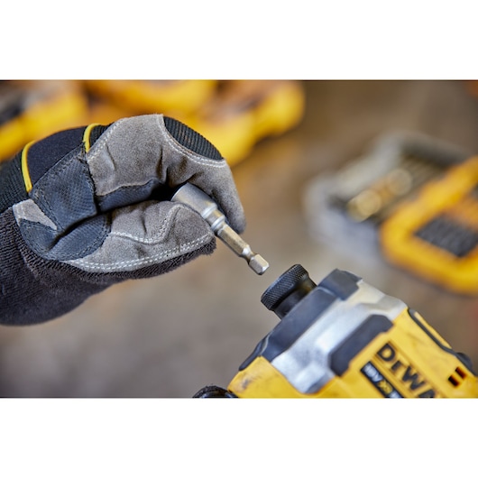 A gloved hand holding a metal screwdriver bit, about to insert it into a yellow DEWALT power tool. Tool bits and yellow
