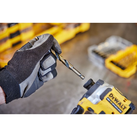 A gloved hand holding a DEWALT drill bit, preparing to attach it to a DEWALT power tool. In the background, yellow DEWALT