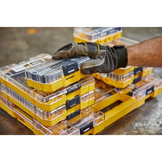 Gloved hand stacking yellow DEWALT storage boxes with clear lids and black clasps on a workshop floor.