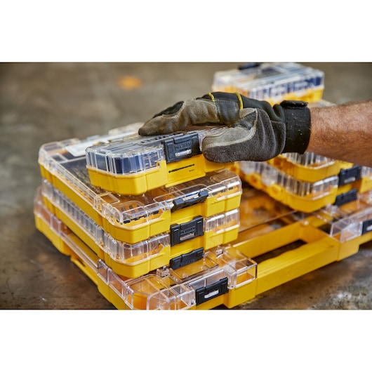Stacked yellow DEWALT tool cases with clear lids. Gloved hand is lifting the top case in a workshop setting.