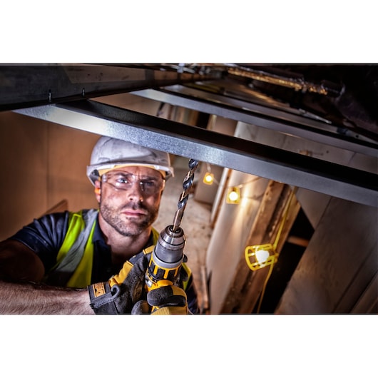 A construction worker wearing safety gear uses a DEWALT drill to work on a metal beam, with industrial lighting visible in