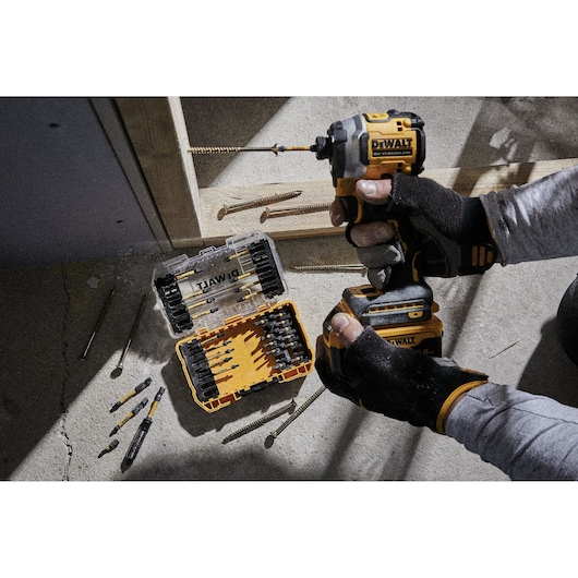 A person with gloves is using a DEWALT cordless impact driver, and a DEWALT bit set in an open yellow and black case is