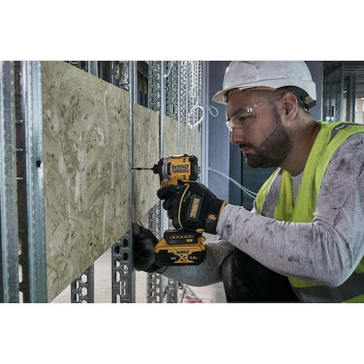 A construction worker wearing a safety helmet and hi-vis vest is using a DEWALT cordless drill to fasten a wooden board onto