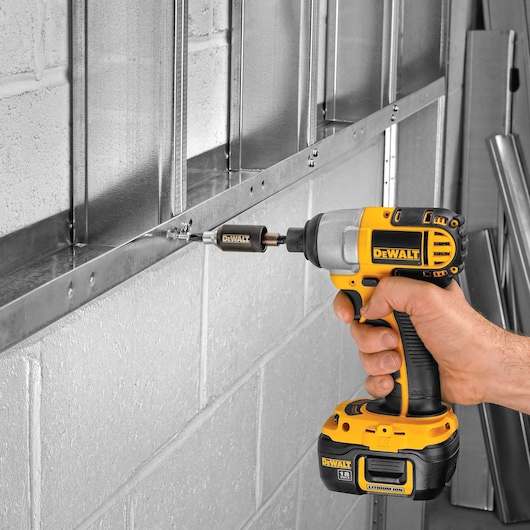 A person is using a DEWALT cordless power drill to fasten a screw into a metal frame attached to a cement block wall.