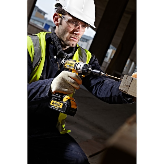 Worker uses a DEWALT cordless drill on wood at a construction site. Safety gear and hi-vis vest visible.