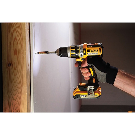 A DEWALT cordless drill is being used to drive a screw into a wall, held by a person wearing a work glove.