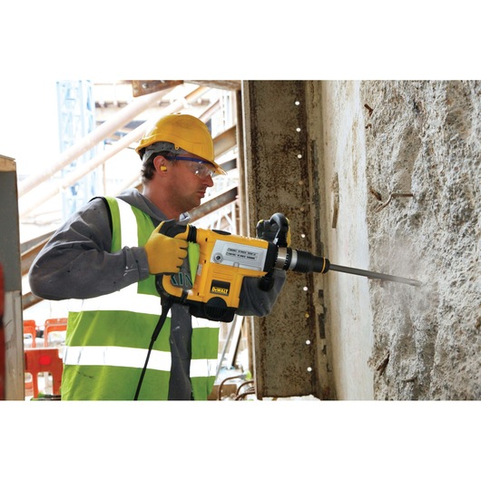 A construction worker wearing a yellow hard hat and high-visibility vest using a DEWALT power tool to drill or break into a concrete wall at a construction site.