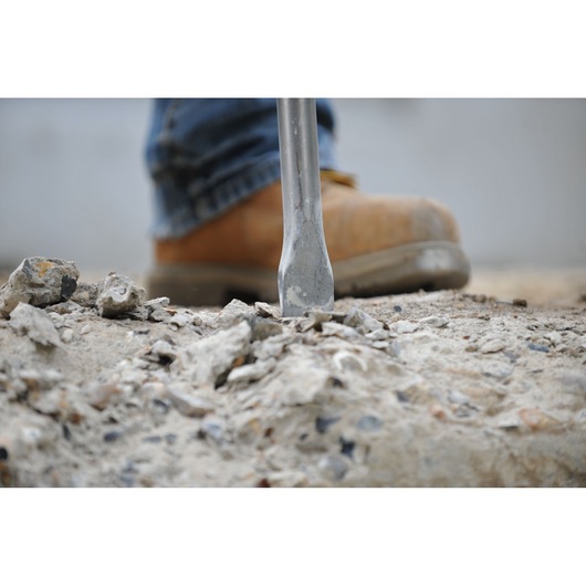 Close-up of a metal chisel breaking concrete, with a person wearing a brown work boot in the background.