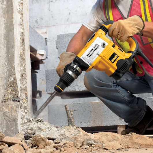 Close-up of a person wearing work gloves and a safety vest using a yellow DEWALT rotary hammer drill to break concrete at a construction site.