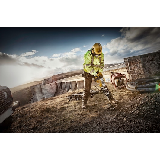 A construction worker wearing a yellow hard hat and high-visibility jacket uses a DEWALT jackhammer to break ground outdoors