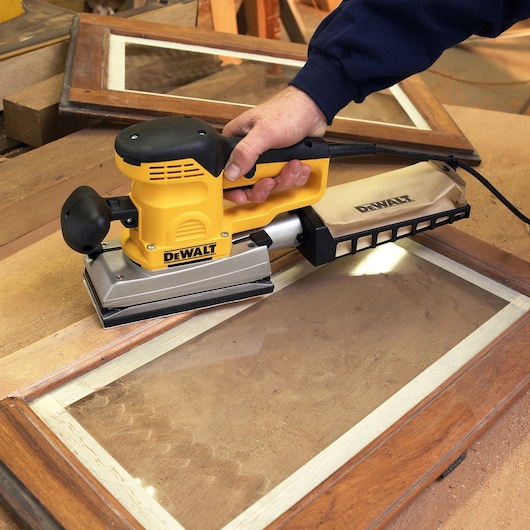 A person using a DEWALT electric sander to sand a wooden window frame. The tool is yellow and black, and the person is holding it with one hand. The workspace includes other wooden materials and tools.
