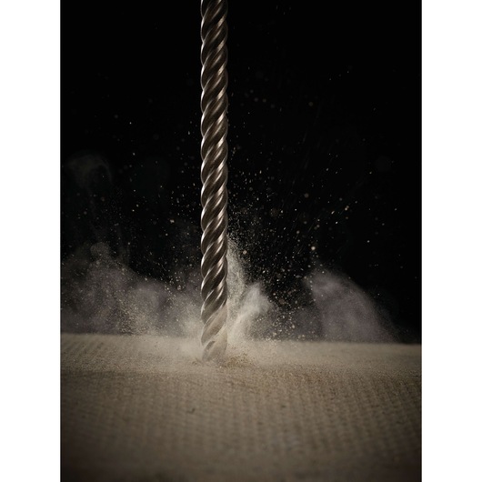 A close-up shot of a metal drill bit penetrating a dusty surface, with particles and dust dispersing around the tip.