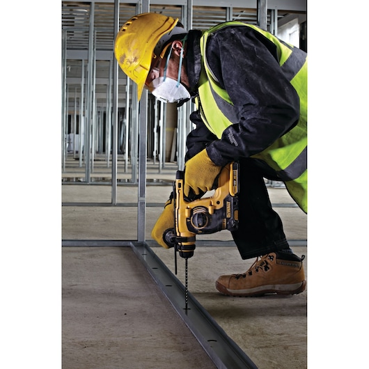 A construction worker wearing a yellow safety helmet, high-visibility vest, gloves, and a face mask is using a DEWALT cordless rotary hammer drill to drill into a metal frame on a construction site.