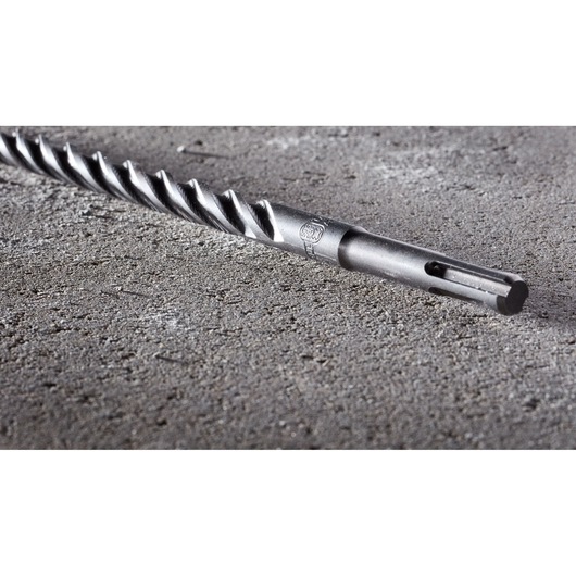 Close-up view of a DEWALT DT8931 SDS drill bit lying on a rough concrete surface, showing the spiral design and fitting end.
