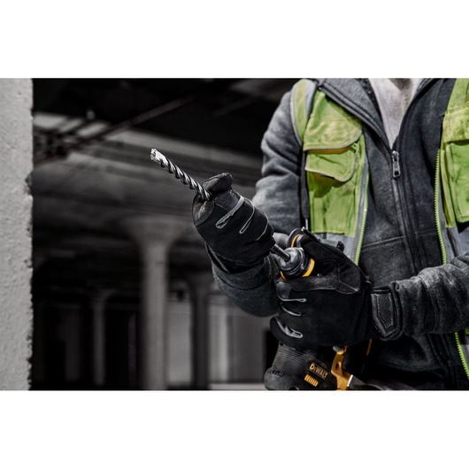 A person wearing black gloves and a safety vest is inserting a DEWALT drill bit into a power drill in an industrial setting.