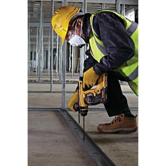 A construction worker wearing a yellow hard hat, high-visibility vest, gloves, and a protective mask is kneeling on the floor while using a DEWALT rotary hammer drill to drill holes into a metal frame in an unfinished building interior.