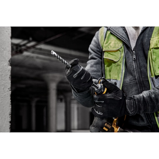 A person wearing work gloves and a green safety vest is holding a DEWALT drill with a spiral drill bit attached, in an industrial indoor setting.