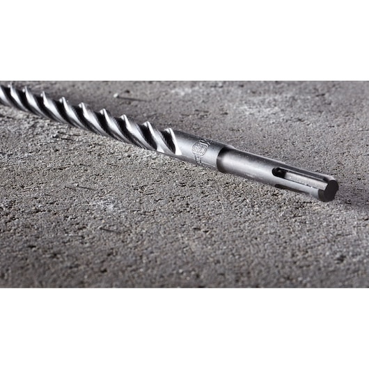 A close-up image of a DEWALT SDS drill bit with a spiral design, lying on a rough concrete surface.