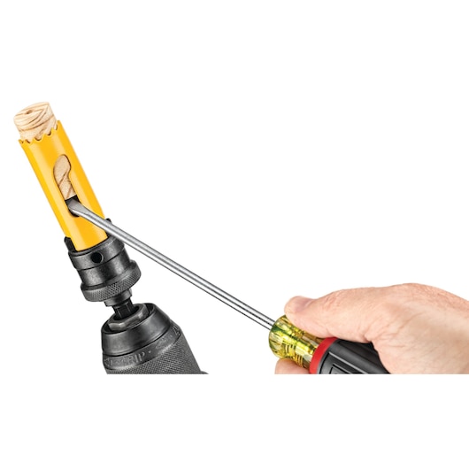 A close-up of a hand using a screwdriver to remove wood debris from a yellow DEWALT hole saw attached to a power drill.