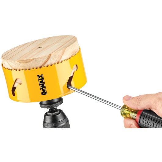 A DEWALT yellow hole saw attached to a drill, with a piece of wood inside. A hand using a screwdriver is removing or adjusting the wood from the hole saw.