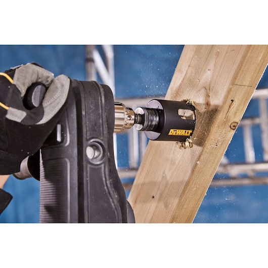 A close-up of a gloved hand operating a drill fitted with a DEWALT hole saw, cutting through a wooden beam. Sawdust is