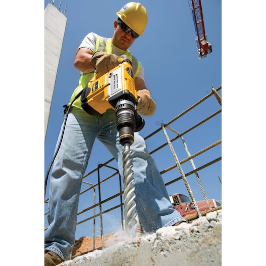 A construction worker using a yellow DEWALT rotary hammer drill to drill into concrete at a building site. The worker is wearing protective gloves, jeans, a high-visibility vest, and a yellow safety helmet. The background shows rebar, a crane, and clear blue sky.