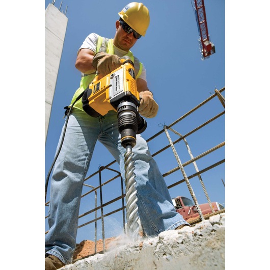 A construction worker using a DEWALT rotary hammer drill to drill into concrete at a building site. The worker is wearing a safety helmet, gloves, and high-visibility vest.