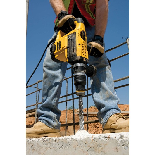 A person using a DEWALT rotary hammer drill with a long drill bit to drill into concrete at a construction site. The person is wearing gloves, work boots, jeans, and a safety vest.