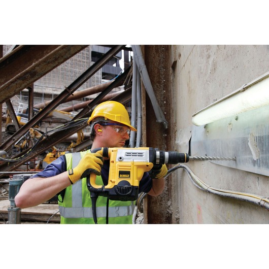 A construction worker wearing a yellow hard hat, ear protection, and high-visibility vest is using a yellow DEWALT rotary hammer drill (SKU: DT9409) to drill into a concrete wall at an industrial site.