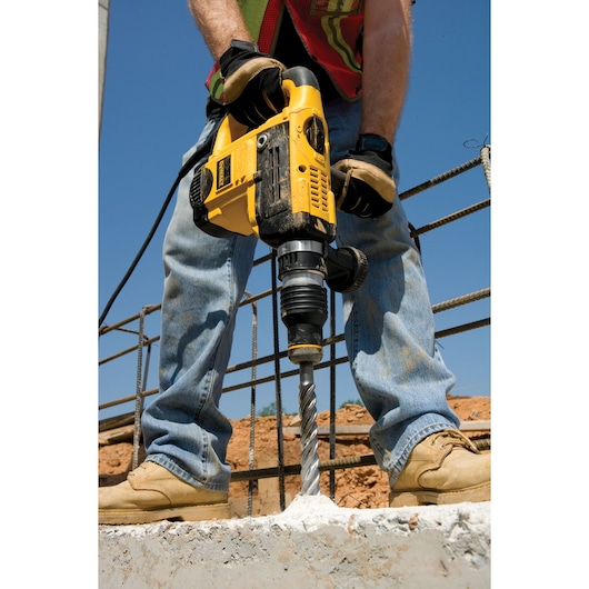 A person wearing work gloves, jeans, boots, and a safety vest is using a yellow DEWALT power drill to bore into a concrete surface at a construction site with metal rebar visible in the background.